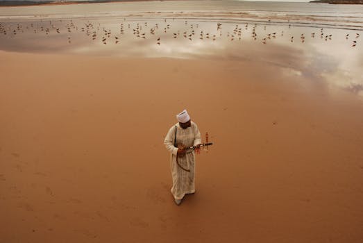 A man in traditional Moroccan clothing plays an instrument on a serene beach.