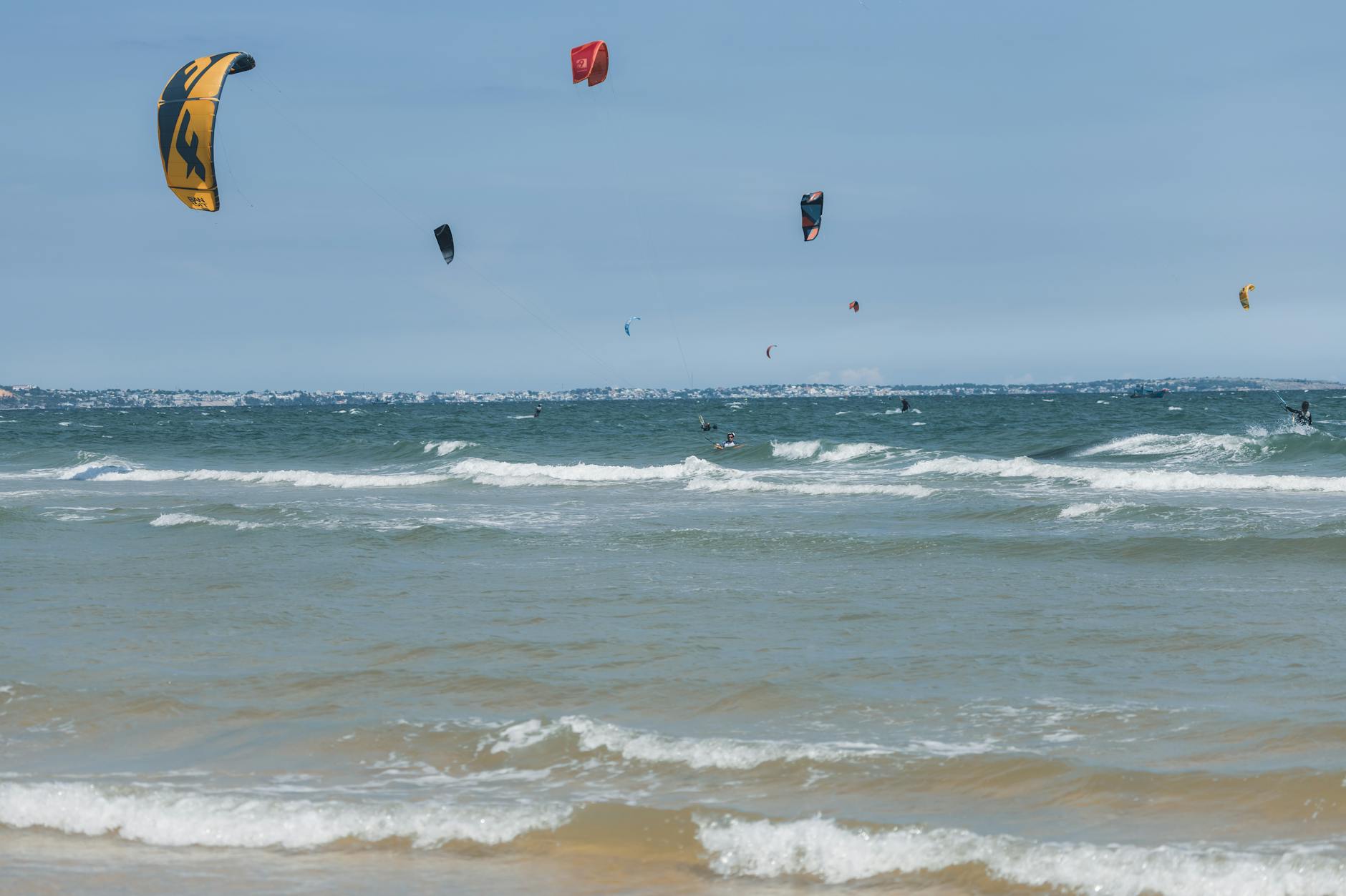 Kitesurfers skillfully riding waves under a clear blue sky with colorful kites.
