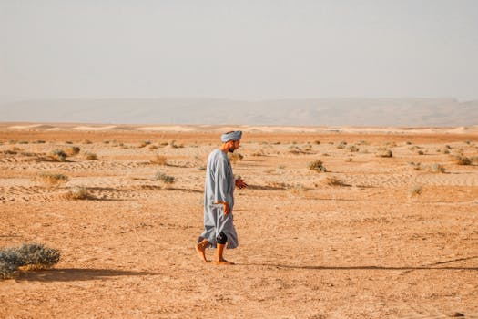 A man in a turban walks barefoot across the sunlit Sahara Desert landscape near Tagounite, Morocco.