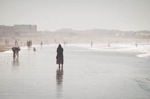 Back view of anonymous female in outerwear standing on icy lake with slippery surface near people walking in town surrounded by mountains in wintertime