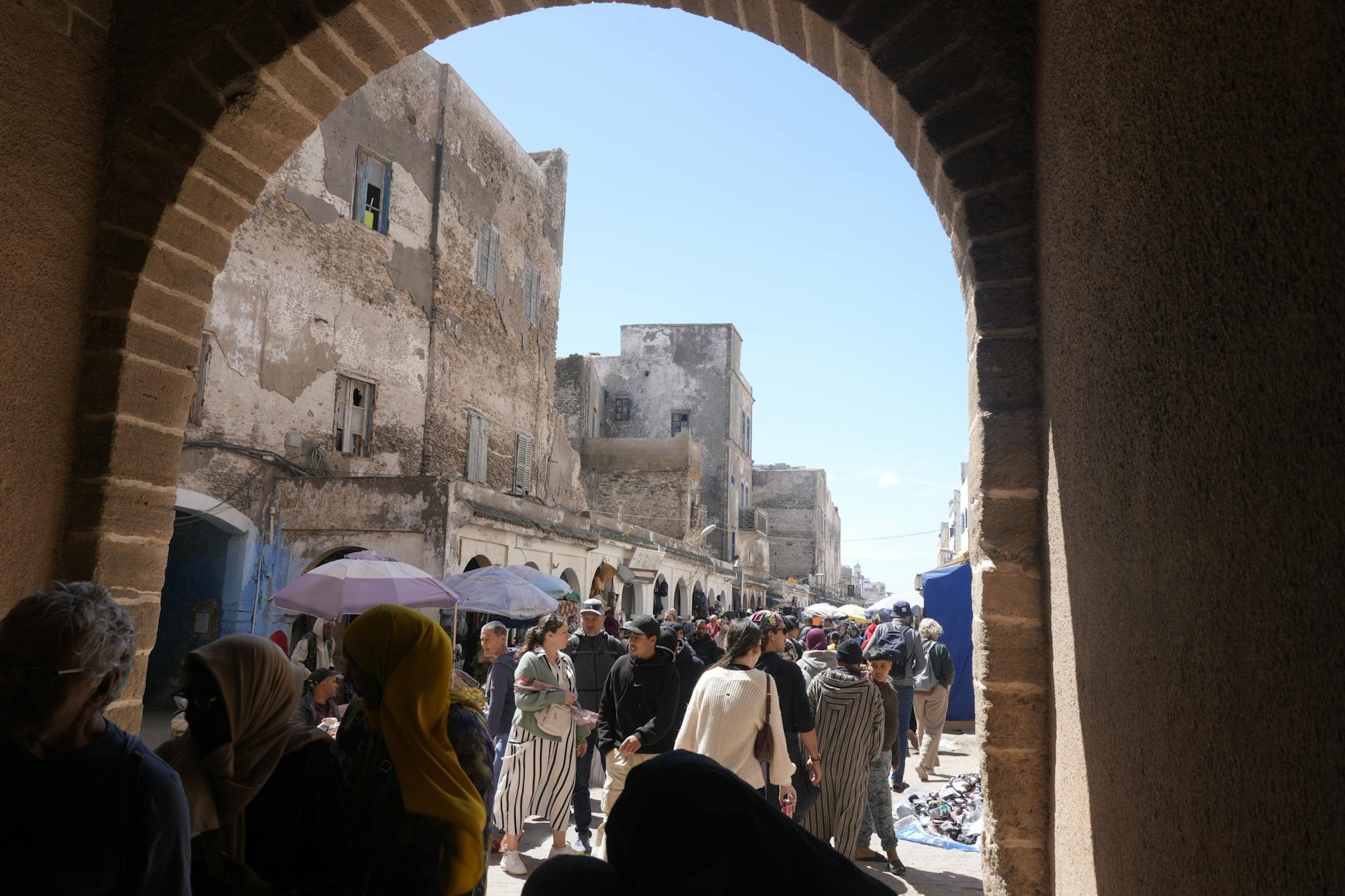 Bustling market scene in Essaouira, Morocco, with people and historical architecture under a bright sky.